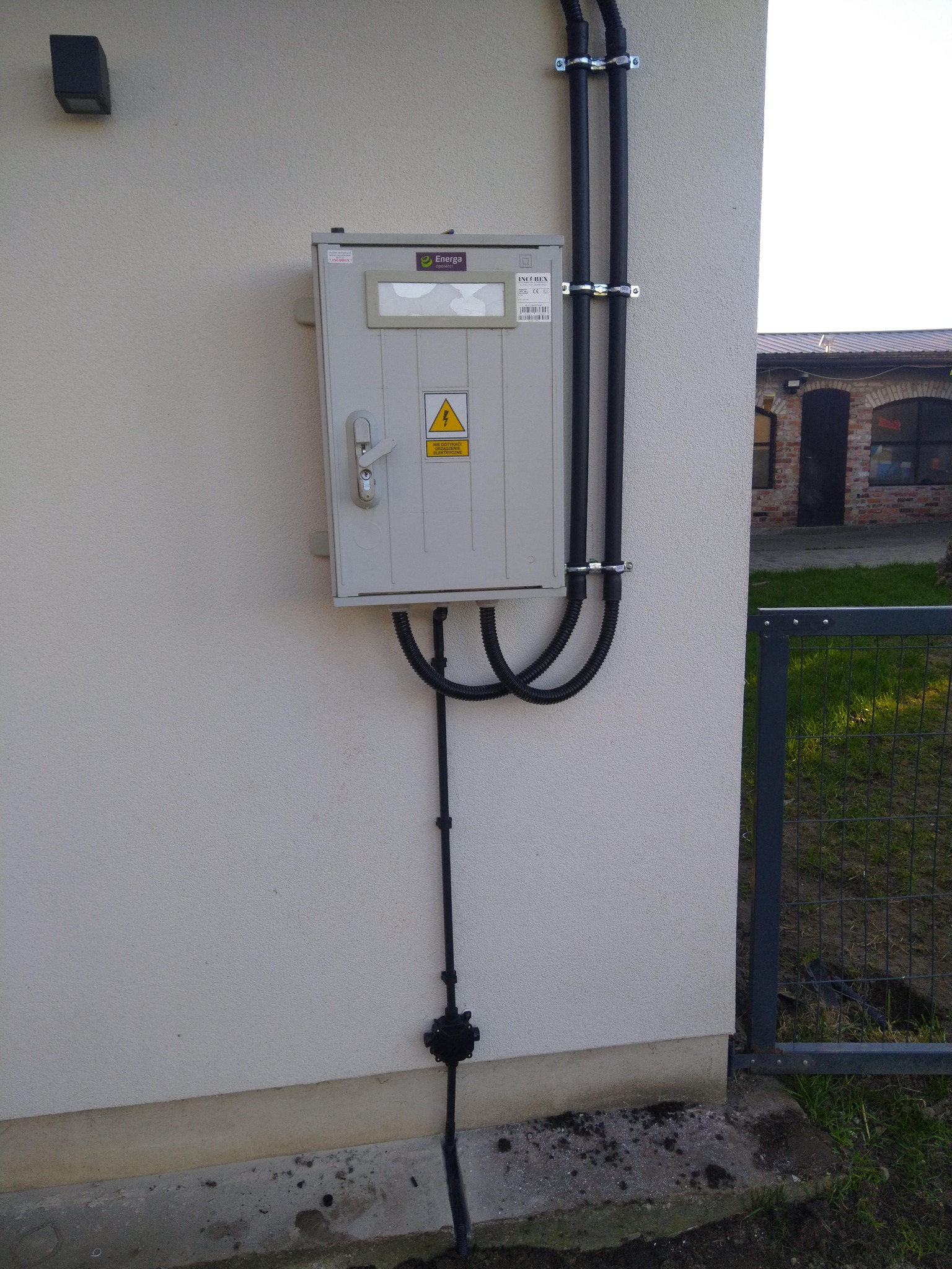 Szara skrzynka elektryczna z symbolem ostrzegawczym zamontowana na elewacji budynku, z widocznymi przewodami w peszlach przymocowanymi do ściany oraz uziemieniem.
