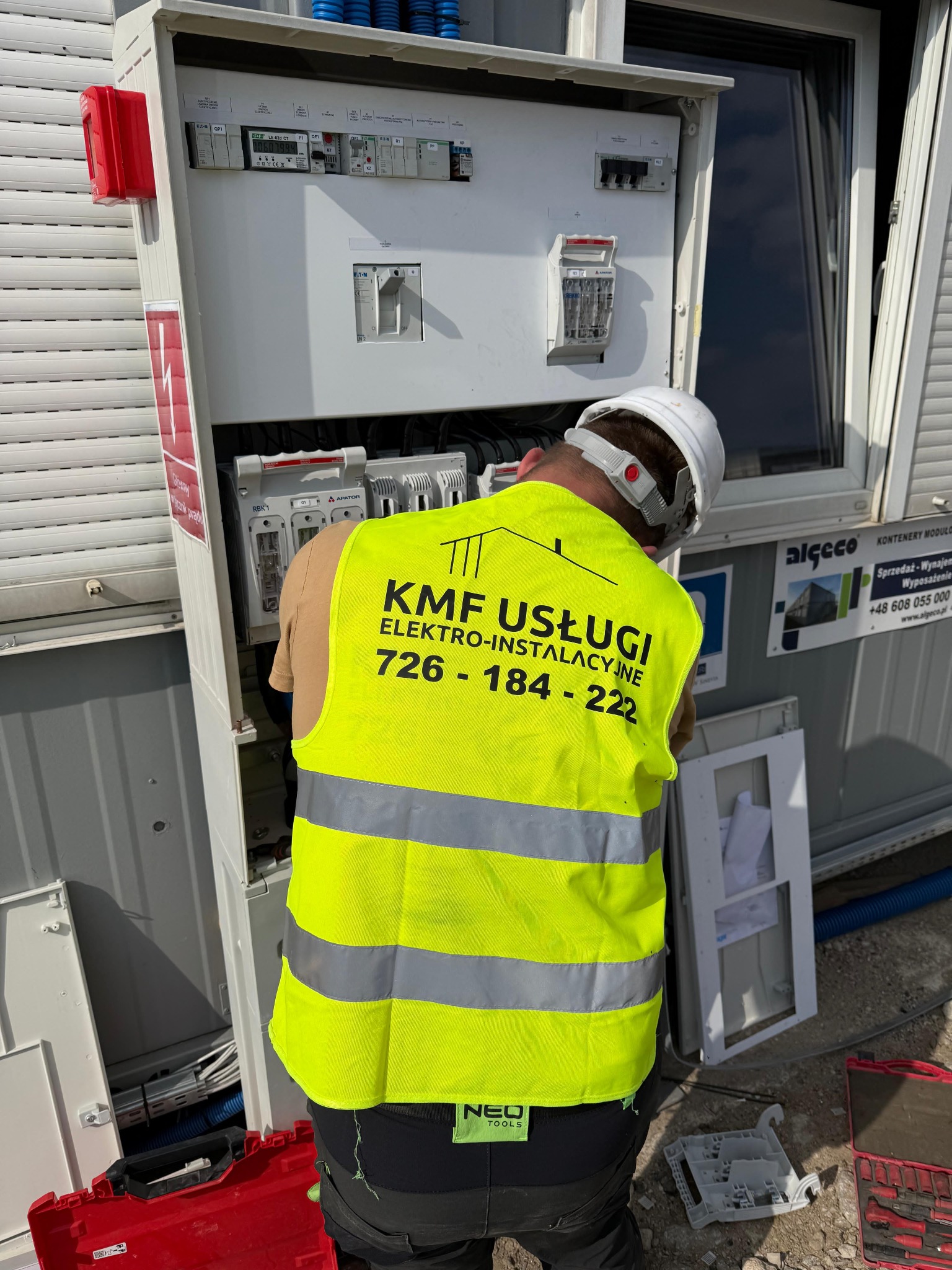Elektryk w żółtej kamizelce z napisem 'KMF Usługi Elektro-Instalacyjne' pracuje przy skrzynce elektrycznej w kontenerze modułowym, widoczne zabezpieczenia i przewody.