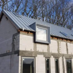 Dom w stanie surowym otwartym z szarym dachem z blachy trapezowej i oknami dachowymi, widoczne drzewa w tle.