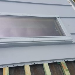 asfasfadfgd - Montaż okna dachowego w pokryciu z blachy na drewnianej konstrukcji, widoczne elementy mocujące i fragment ubrania roboczego