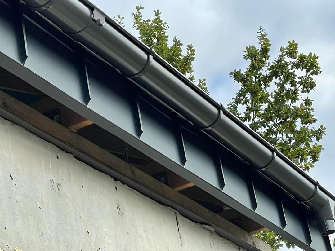 Czarny, metalowy system rynnowy i blaszane pokrycie dachu na tle betonowej ściany i zielonego drzewa.