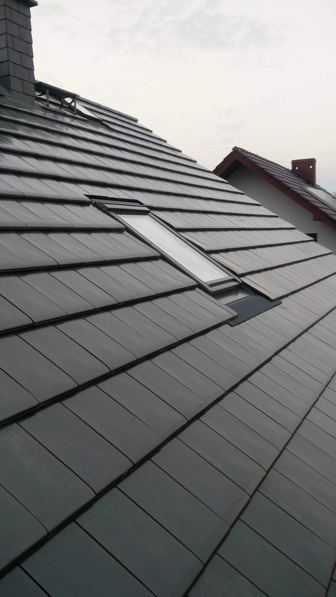 Nowoczesny dach z ciemnej dachówki ceramicznej z oknami dachowymi, widok z dołu na tle pochmurnego nieba.