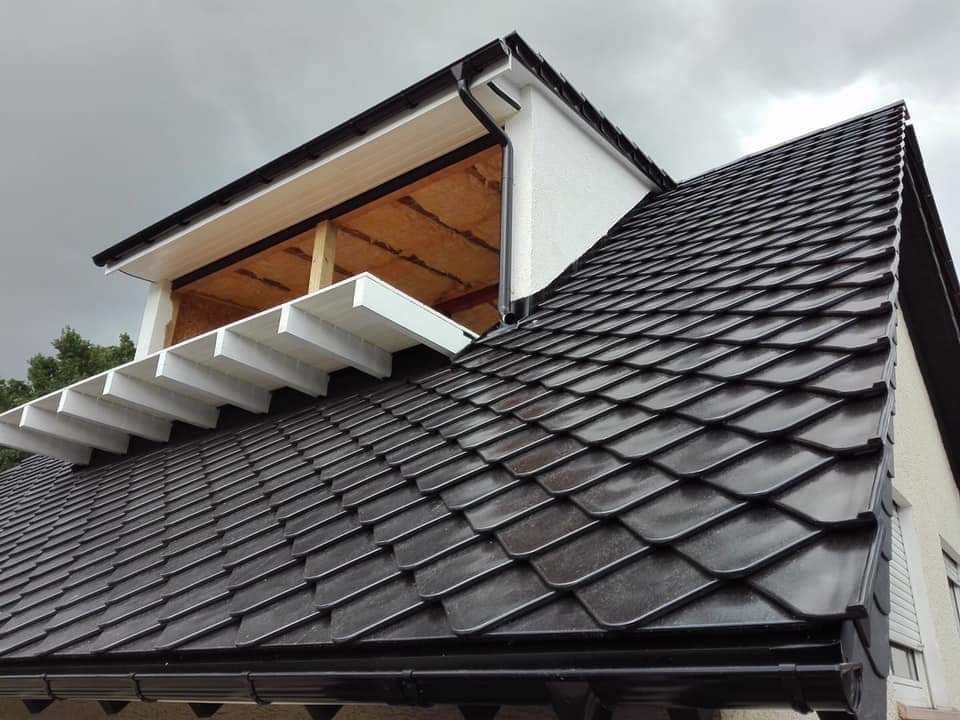Dach pokryty ciemną, łuskową dachówką, widoczna konstrukcja lukarny z ociepleniem, pochmurne niebo w tle.