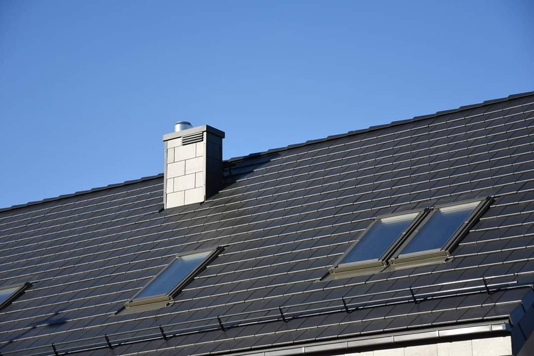 Ciemnoszary dach z dachówki ceramicznej z trzema oknami dachowymi i kominem z szarej cegły na tle błękitnego nieba.