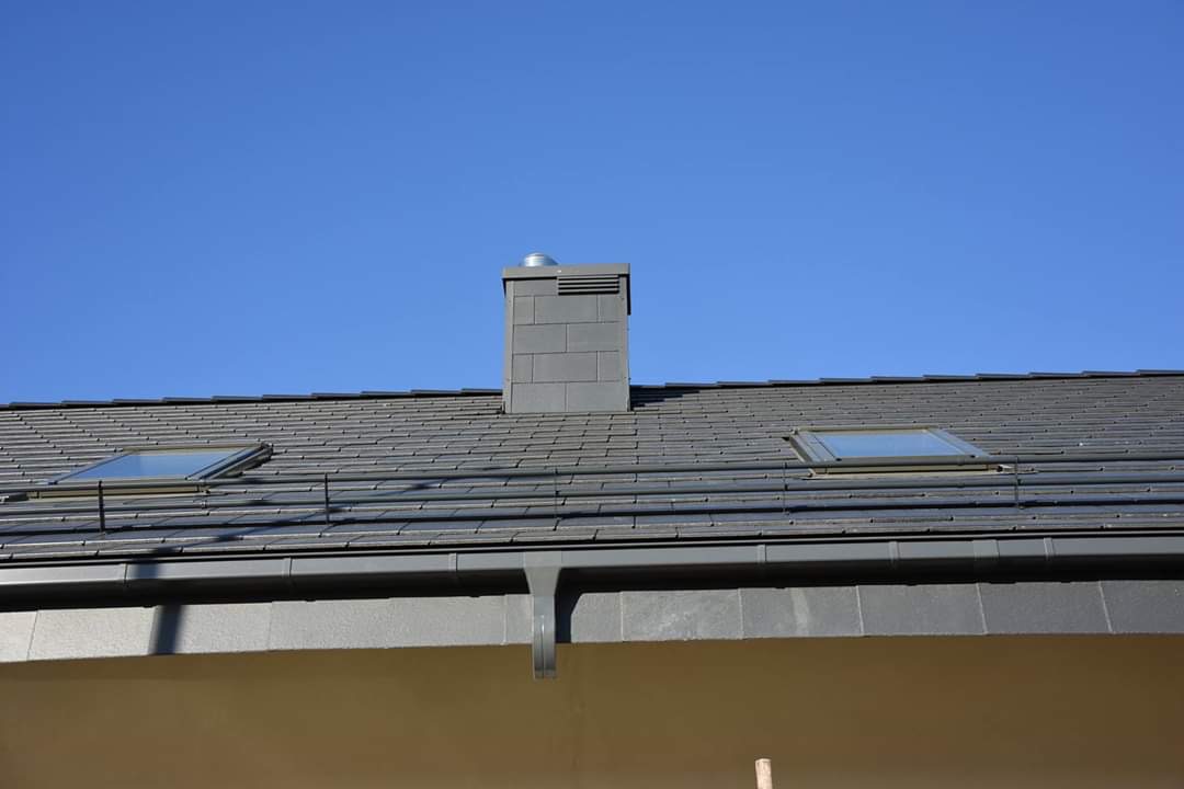 Nowoczesny dach z ciemnoszarej dachówki ceramicznej, dwa okna dachowe i komin z szarej cegły na tle błękitnego nieba.