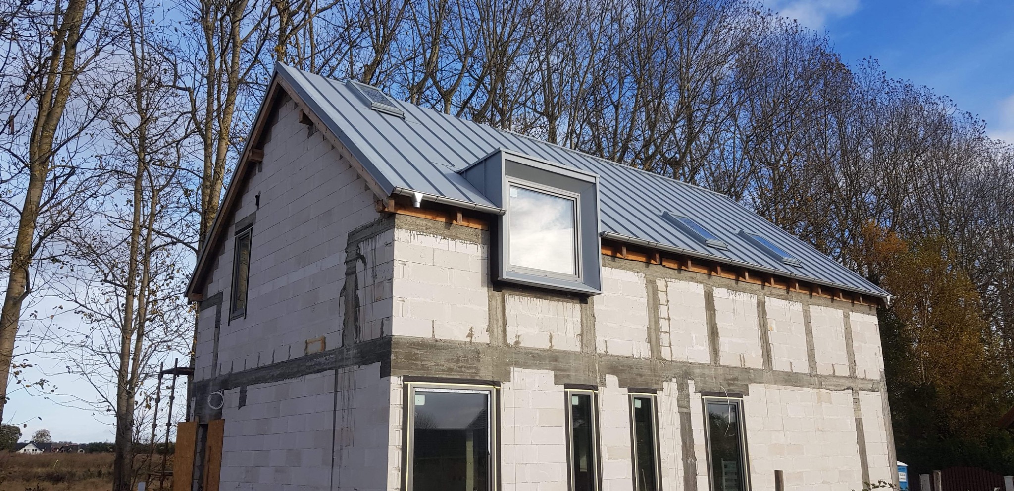 Dom w stanie surowym otwartym z szarym dachem z blachy trapezowej i oknami dachowymi, widoczne drzewa w tle.