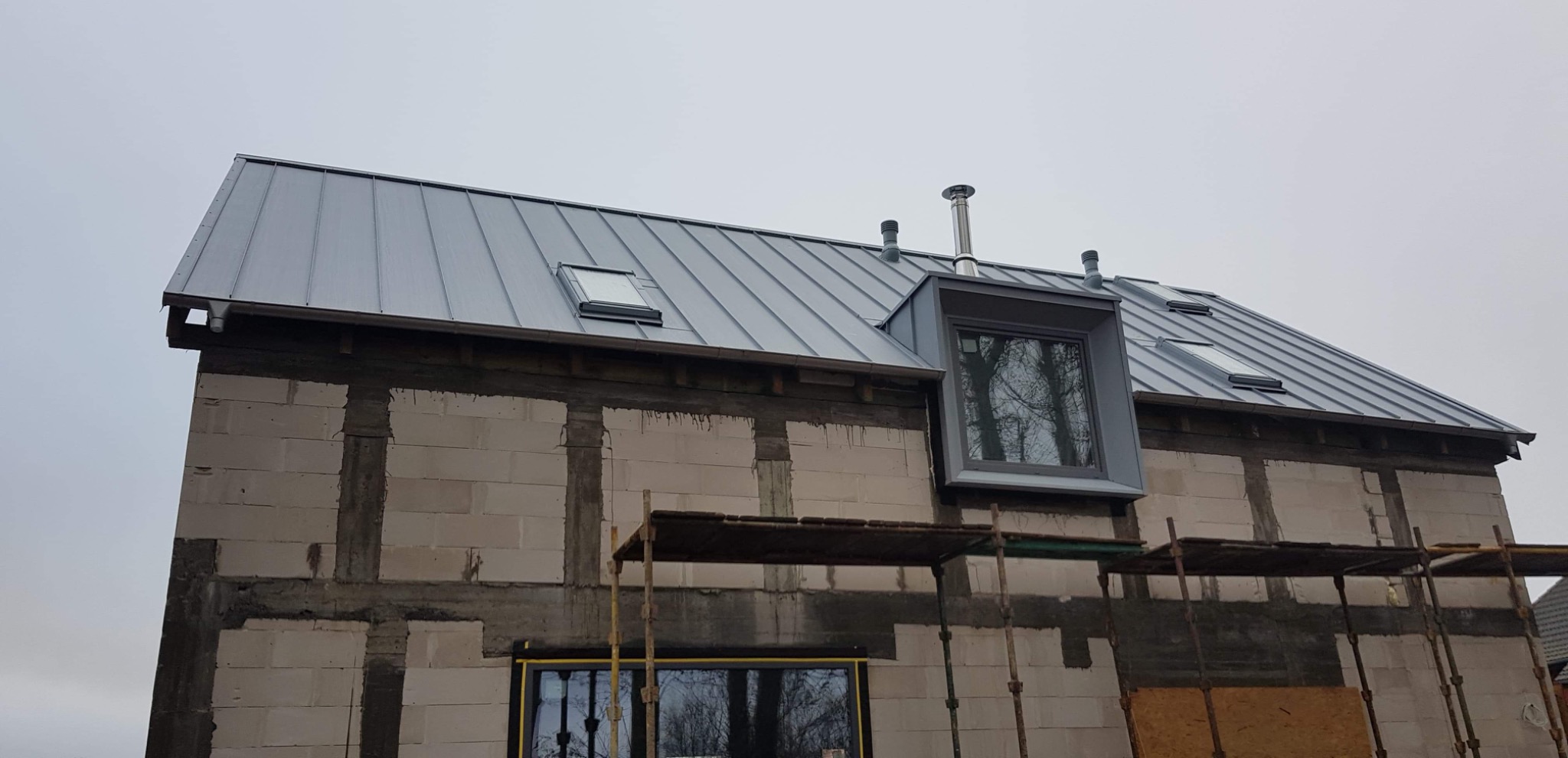 Szary dach z blachy na surowym budynku z oknami dachowymi i kominem, widoczne rusztowanie.