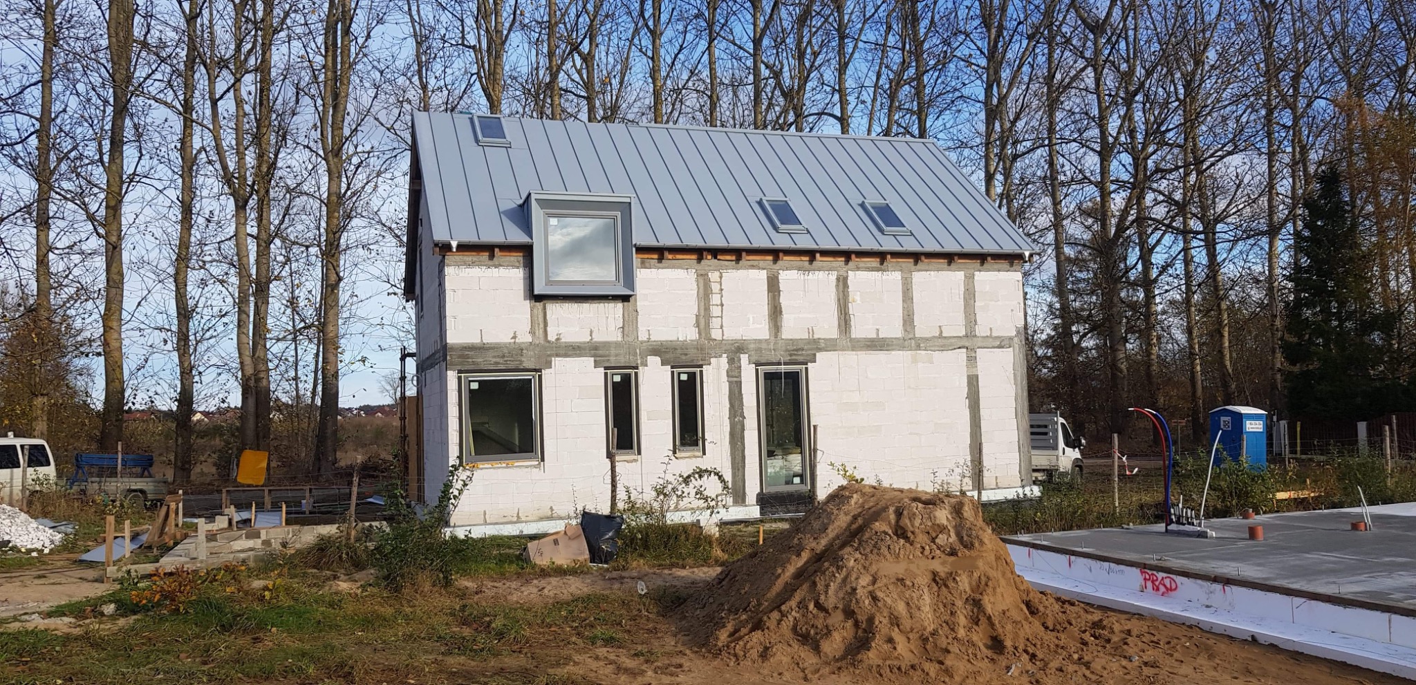Budowa domu jednorodzinnego w trakcie realizacji, widoczna surowa bryła budynku z szarego bloczka, z zamontowanymi oknami i nowym, szarym dachem z lukarnami, na pierwszym planie hałda ziemi.