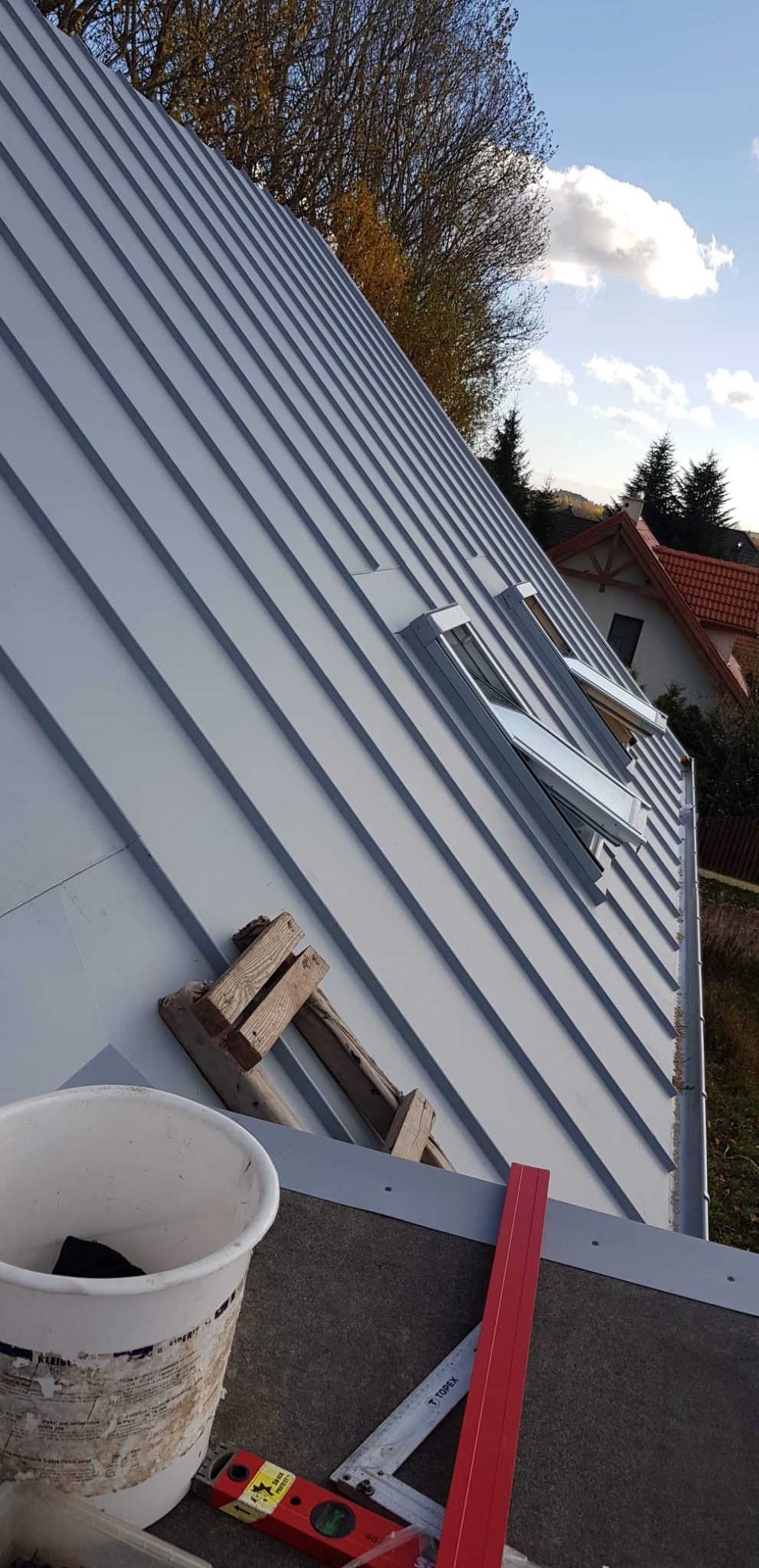 Nowy, blaszany dach w kolorze szarym z dwoma oknami dachowymi, widoczne narzędzia pracy: poziomica, miarka, wiadro oraz fragment drabiny dekarskiej.
