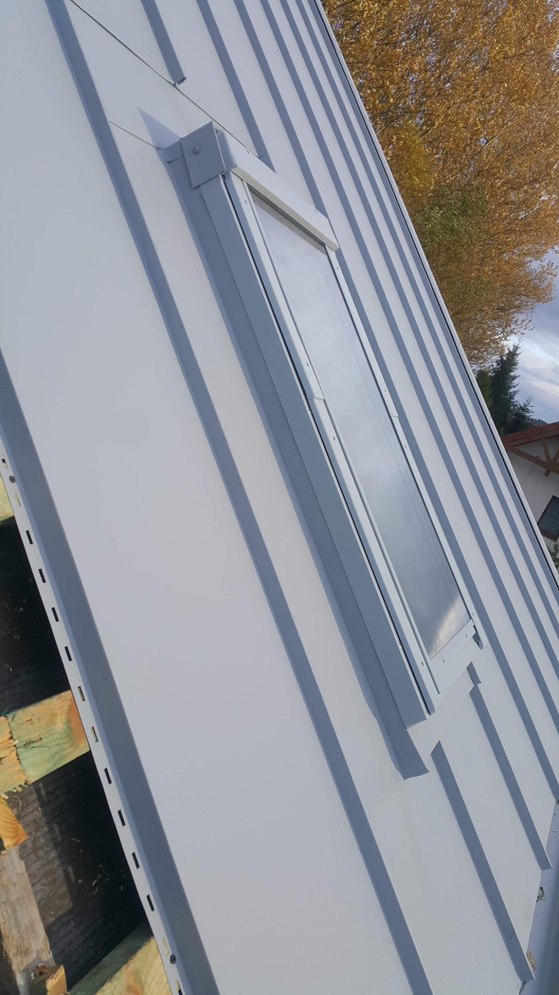 Szary dach z blachy trapezowej z oknem dachowym, widoczne elementy drewnianej konstrukcji w dolnej części kadru.