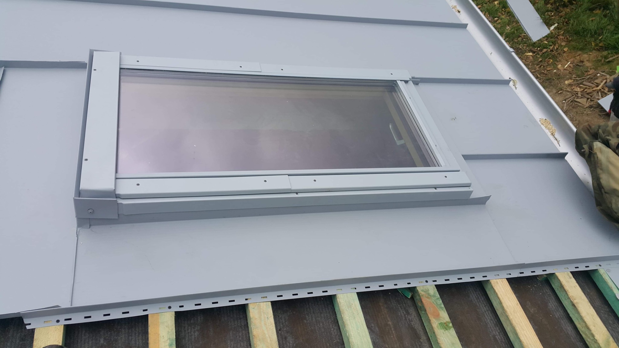 Montaż okna dachowego w pokryciu z blachy na drewnianej konstrukcji, widoczne elementy mocujące i fragment ubrania roboczego