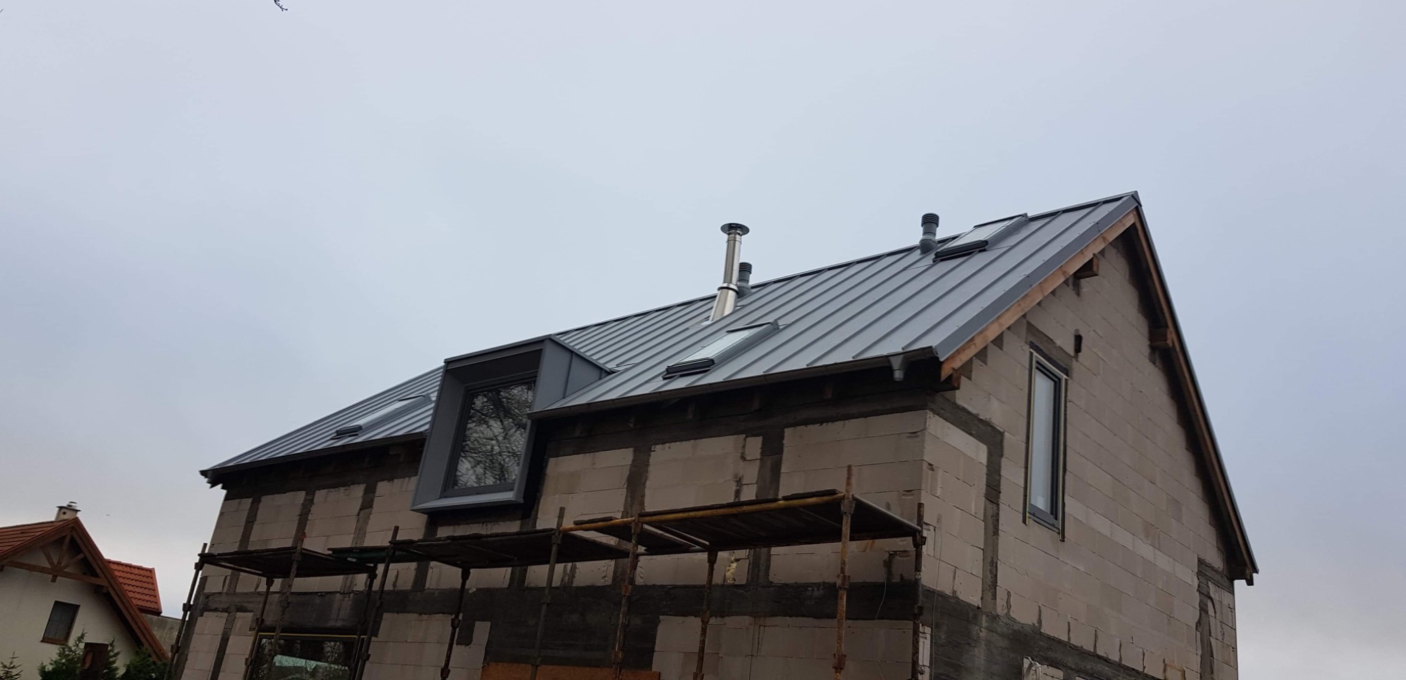 Dom w trakcie budowy z nowym, metalowym dachem z oknami dachowymi i kominem, widoczne rusztowania.