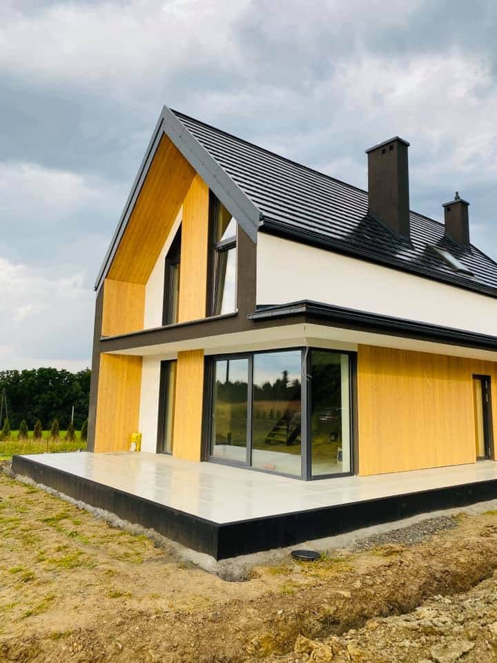 Nowoczesny dom jednorodzinny z ciemną, metalową dachówką, drewnianą elewacją i dużymi przesuwnymi oknami tarasowymi, częściowo wykończony taras.