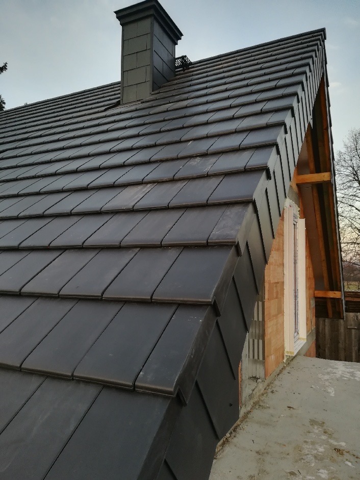 Nowy, ciemny dach z dachówki ceramicznej, widoczny komin w tym samym kolorze, fragment ściany szczytowej z oknem w trakcie budowy.
