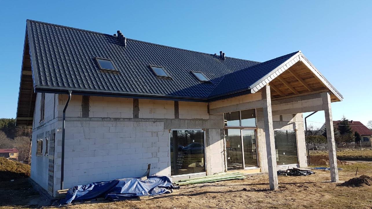 Surowy dom jednorodzinny w trakcie budowy z ciemną dachówką, widoczne okna dachowe, rynny i ściany z betonu komórkowego, na ziemi leżą materiały budowlane.