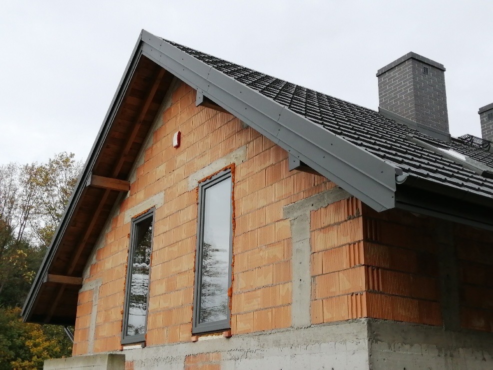 Dom w trakcie budowy z widoczną konstrukcją dachu, oknami w surowym stanie i ceglaną elewacją, na tle pochmurnego nieba i zieleni.