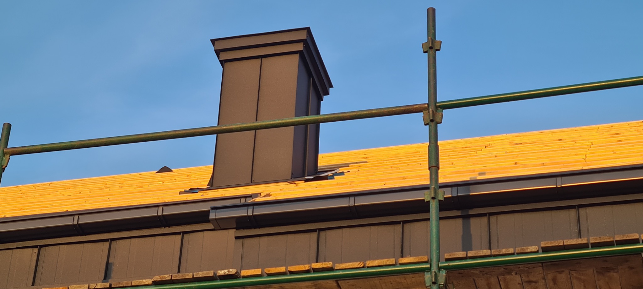 Widok na dach w trakcie montażu z widoczną więźbą, kominem w ciemnym kolorze i fragmentem rusztowania na tle błękitnego nieba.