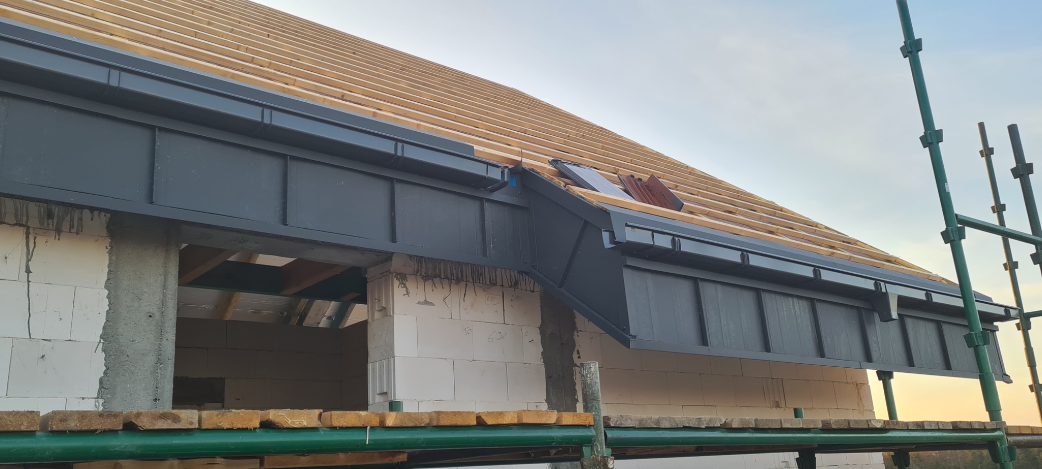 Montaż drewnianej konstrukcji dachu z zamontowanymi, grafitowymi elementami orynnowania na surowym budynku z białego bloczka.