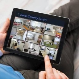 Agnieszka Purczyńska SV-NET - Tablet z aplikacją 'Home Security System' wyświetlającą podgląd z kamer monitoringu w różnych pomieszczeniach domu, trzymany przez osobę wskazującą palcem na ekran.