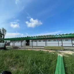 Budowa zadaszenia z zielonych belek na konstrukcji budynku z pustak&oacute;w, widok na plac budowy z trawą i sprzętem budowlanym w tle, błękitne niebo z chmurami.