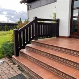 Zewnętrzne schody wejściowe z terakoty z dekoracyjnym wzorem na krawędziach stopni i ciemnobrązową drewnianą barierką, widok na dom z białym tynkiem i panele słoneczne w ogrodzie.