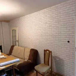 Ściana wykończona tapetą imitującą białą cegłę w pokoju z drewnianą podłogą, widoczny stary fotel, krzesło i stolik z resztkami po pracy.