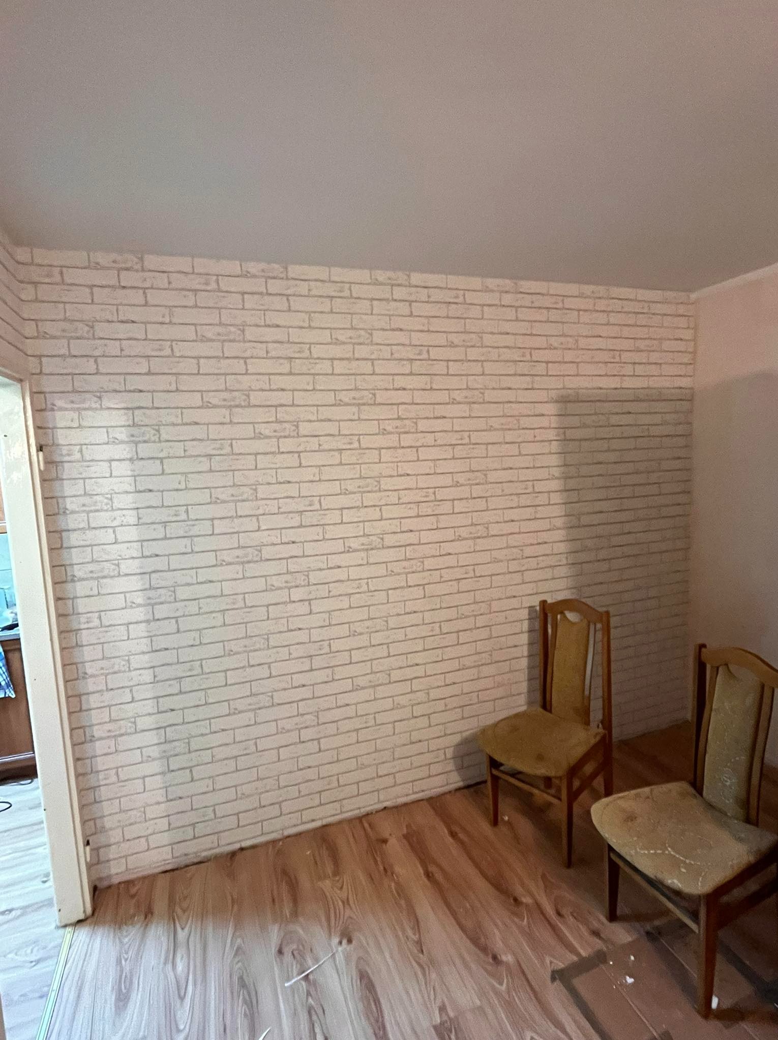 Ściana wykończona tapetą imitującą białą cegłę, z widocznymi dwoma starymi krzesłami i fragmentem parkietu na podłodze.