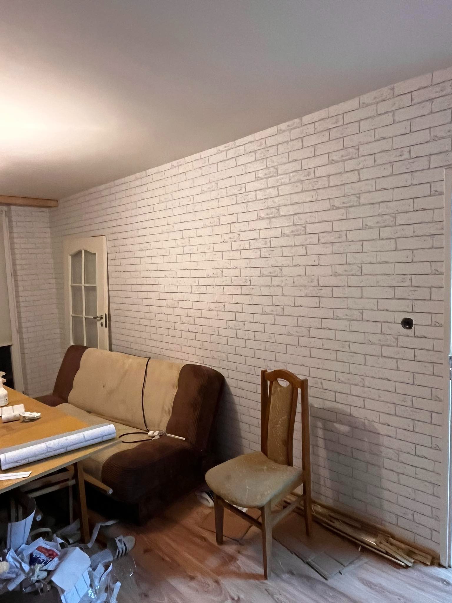 Ściana wykończona tapetą imitującą białą cegłę w pokoju z drewnianą podłogą, widoczny stary fotel, krzesło i stolik z resztkami po pracy.