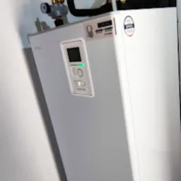 Biała pompa ciepła w rogu pomieszczenia, widoczny zbiornik wyr&oacute;wnawczy i izolowane rury, panel sterowania z zieloną diodą.