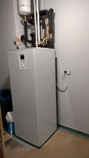 Nowoczesna, szara pompa ciepła w kotłowni, z widocznymi rurami instalacji grzewczej i elementami montażowymi, zainstalowana przy szarej ścianie z wystającą puszką elektryczną.