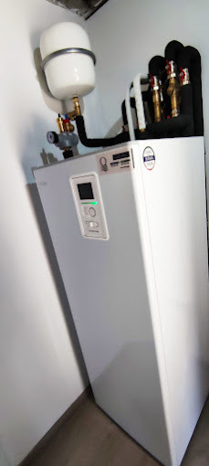 Biała pompa ciepła w rogu pomieszczenia, widoczny zbiornik wyrównawczy i izolowane rury, panel sterowania z zieloną diodą.