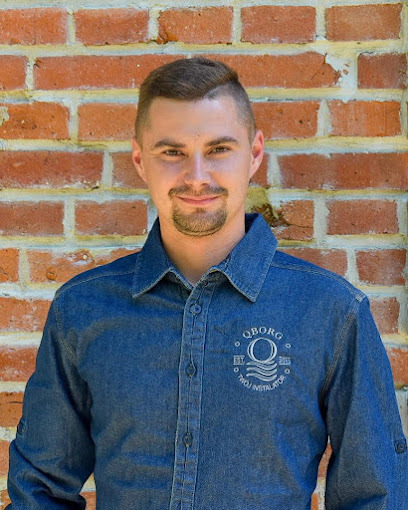 Portret uśmiechniętego mężczyzny w dżinsowej koszuli z logo firmy instalacyjnej na tle ceglanej ściany.