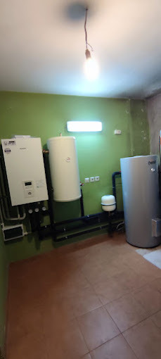 Instalacja grzewcza w pomieszczeniu z zielonymi ścianami i brązową podłogą, widoczne kotły, zbiorniki i rury, oświetlenie sufitowe z żarówką i lampą jarzeniową.