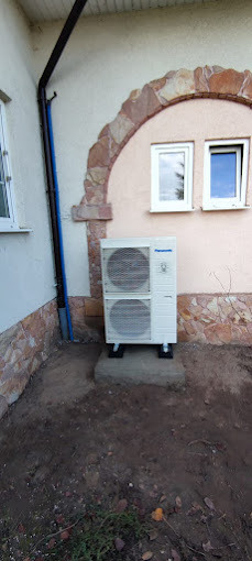 Zewnętrzna jednostka pompy ciepła Panasonic zamontowana na betonowym fundamencie przy ścianie budynku z dekoracyjnym łukiem z kamienia. Widoczne rury odprowadzające skropliny.