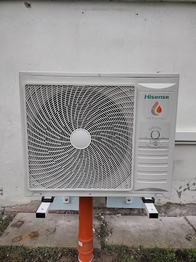 Zewnętrzna jednostka pompy ciepła Hisense Hi-Thermo zamontowana na wspornikach na pomarańczowej rurze, widok z przodu na tle białej ściany budynku.