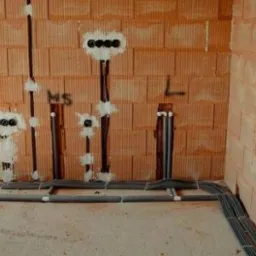 Surowe ściany z cegły z widocznymi przewodami elektrycznymi poprowadzonymi w bruzdach i puszkami pod gniazdka, zabezpieczonymi białym gipsem, widoczne oznaczenia 'MS' i 'L' na ścianie.