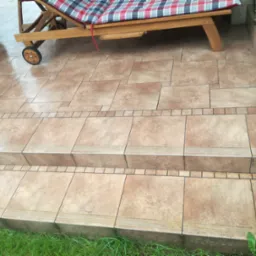 Wyłożony brązowymi płytkami taras z dwoma stopniami, na którym stoi drewniany leżak z materacem w kratę.