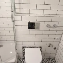 Nowoczesna łazienka z białymi, prostokątnymi płytkami na ścianach, wzorzystą podłogą w odcieniach szarości i czerni, oraz wiszącą miską WC z chromowanym przyciskiem spłukującym.