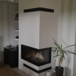 Nowoczesny kominek narożny z czarnym obramowaniem w jasnym salonie w Wyszkowie, obok roślina doniczkowa i dekoracyjne lampiony.