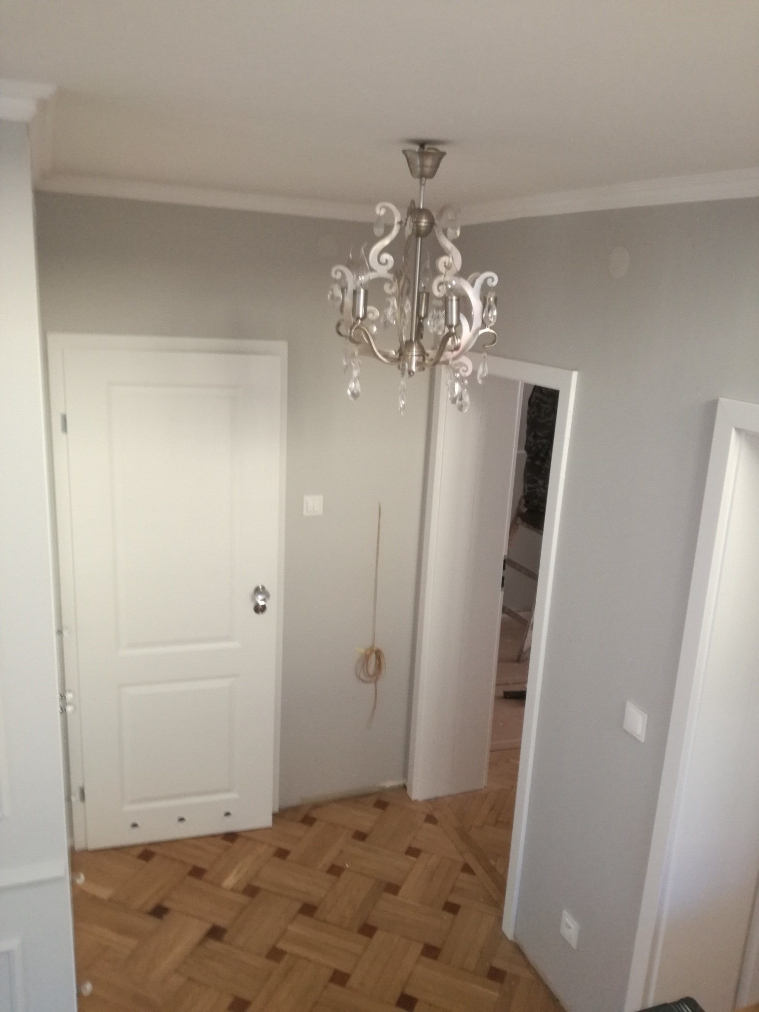 Wnętrze pokoju z parkietem w jodełkę, świeżo pomalowanymi szarymi ścianami, białymi drzwiami z srebrnymi klamkami i ozdobnym żyrandolem w stylu vintage.