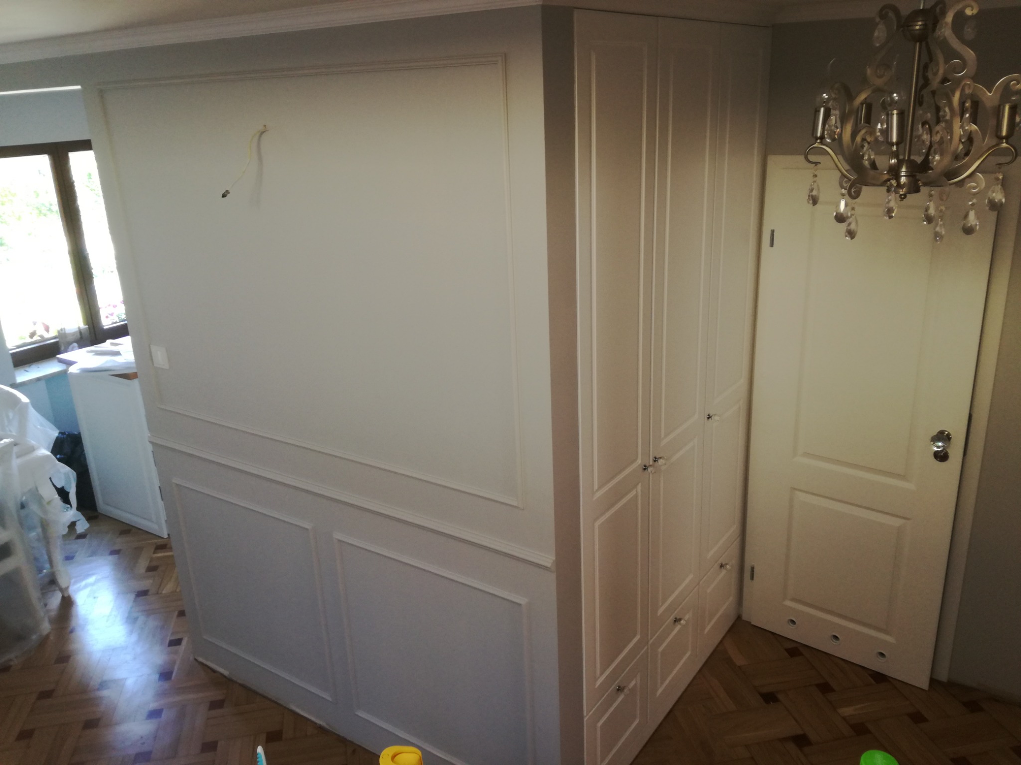 Biała ściana z listwami dekoracyjnymi i wystającymi kablami elektrycznymi, obok biała szafa wnękowa z kryształowymi gałkami, parkiet w jodełkę i ozdobny żyrandol.