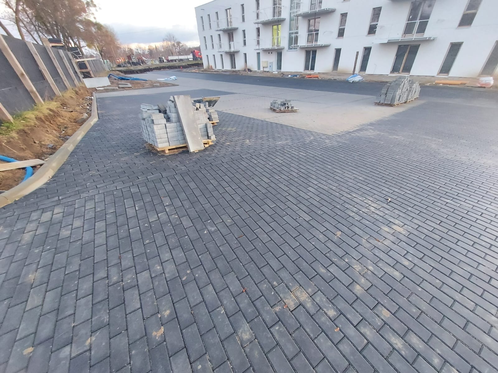 Częściowo ułożona, ciemnoszara nawierzchnia z kostki brukowej w kształcie prostokątów, z paletą kostki i budynkiem mieszkalnym w tle.