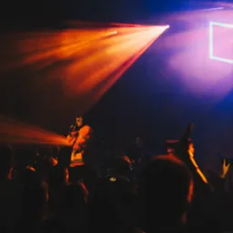 Wokalista w pomarańczowej kurtce śpiewa do mikrofonu na scenie oświetlonej reflektorami, w tle perkusja i gitarzysta, a przed sceną widownia.