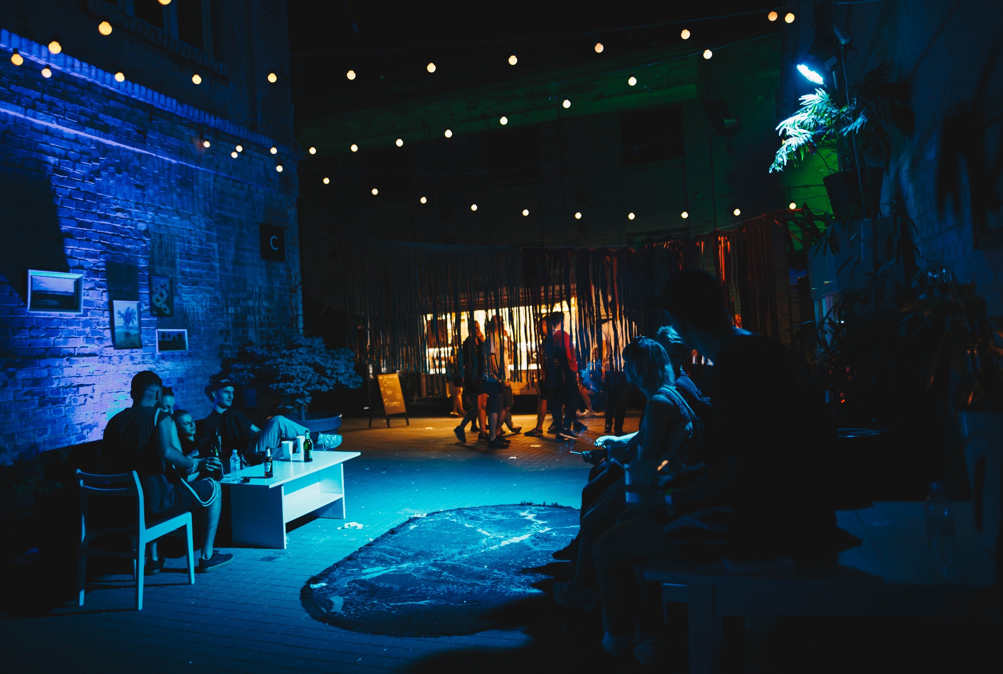 Oświetlona girlandami przestrzeń eventowa z siedzącymi gośćmi przy białym stoliku i przechodzącymi osobami w tle, w chłodnej, niebiesko-zielonej tonacji.