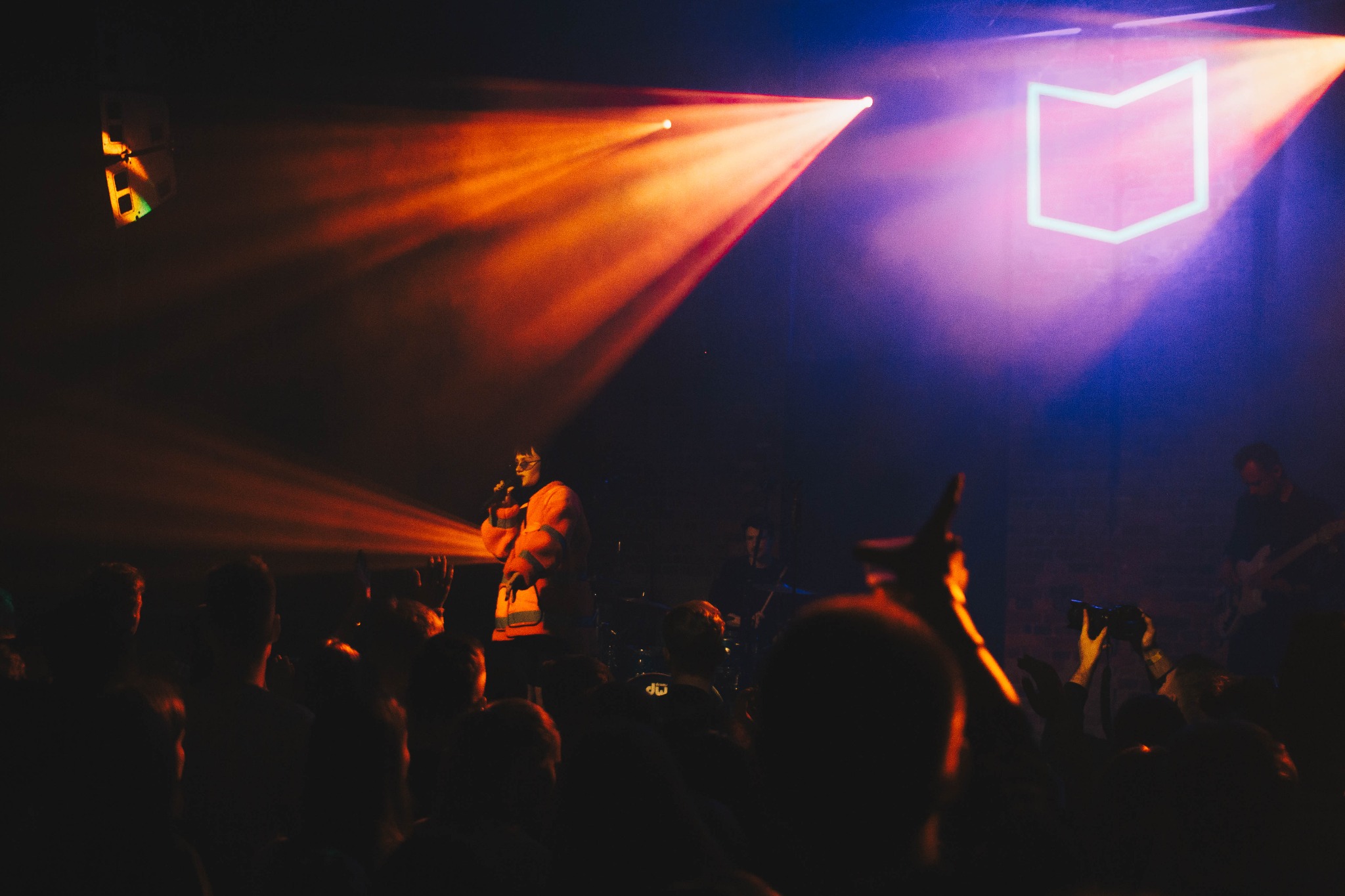 Wokalista w pomarańczowej kurtce śpiewa do mikrofonu na scenie oświetlonej reflektorami, w tle perkusja i gitarzysta, a przed sceną widownia.