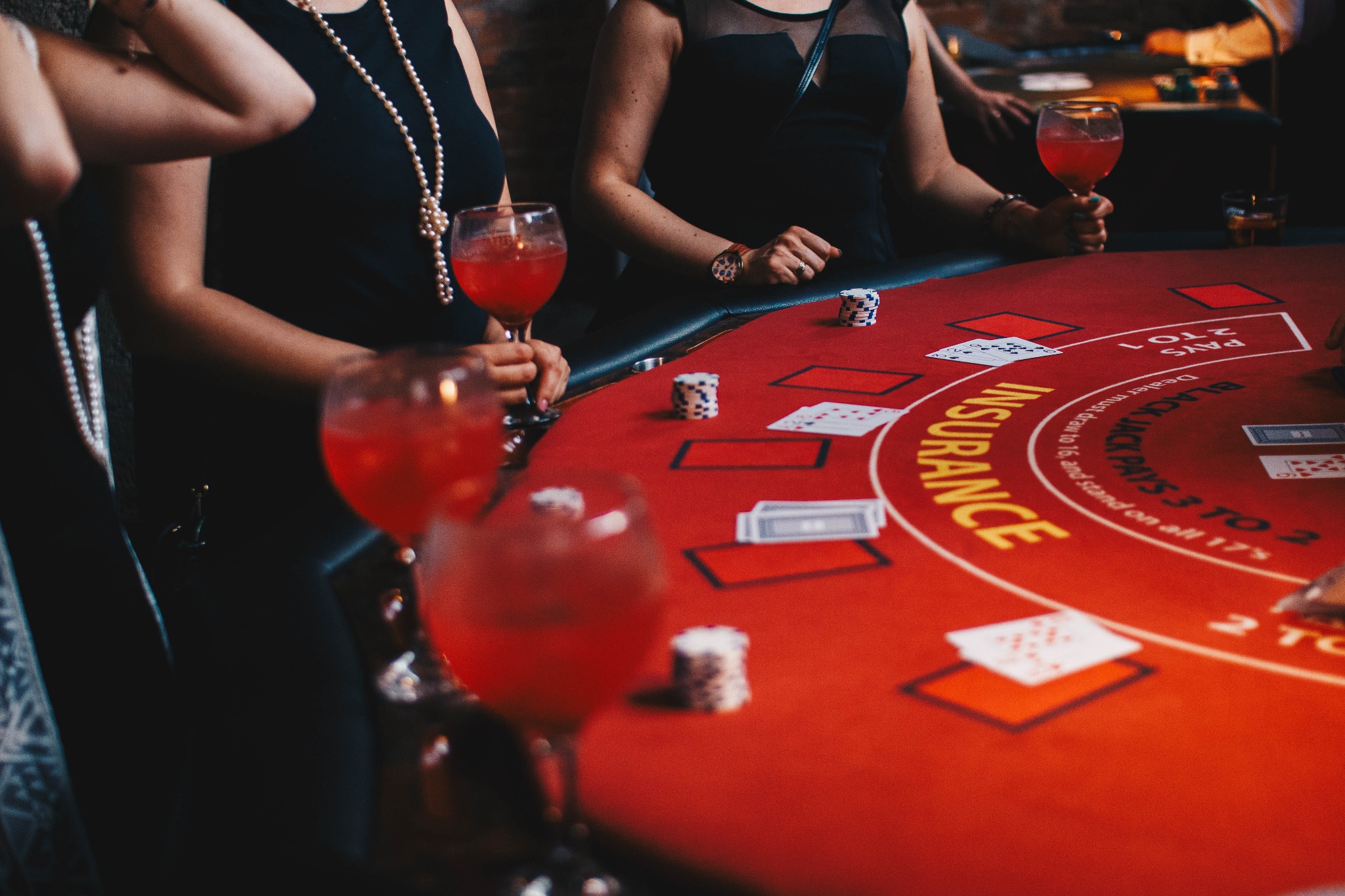 Kobiety w eleganckich sukienkach przy czerwonym stole do blackjacka z żetonami i kartami, trzymające kieliszki z czerwonym napojem.