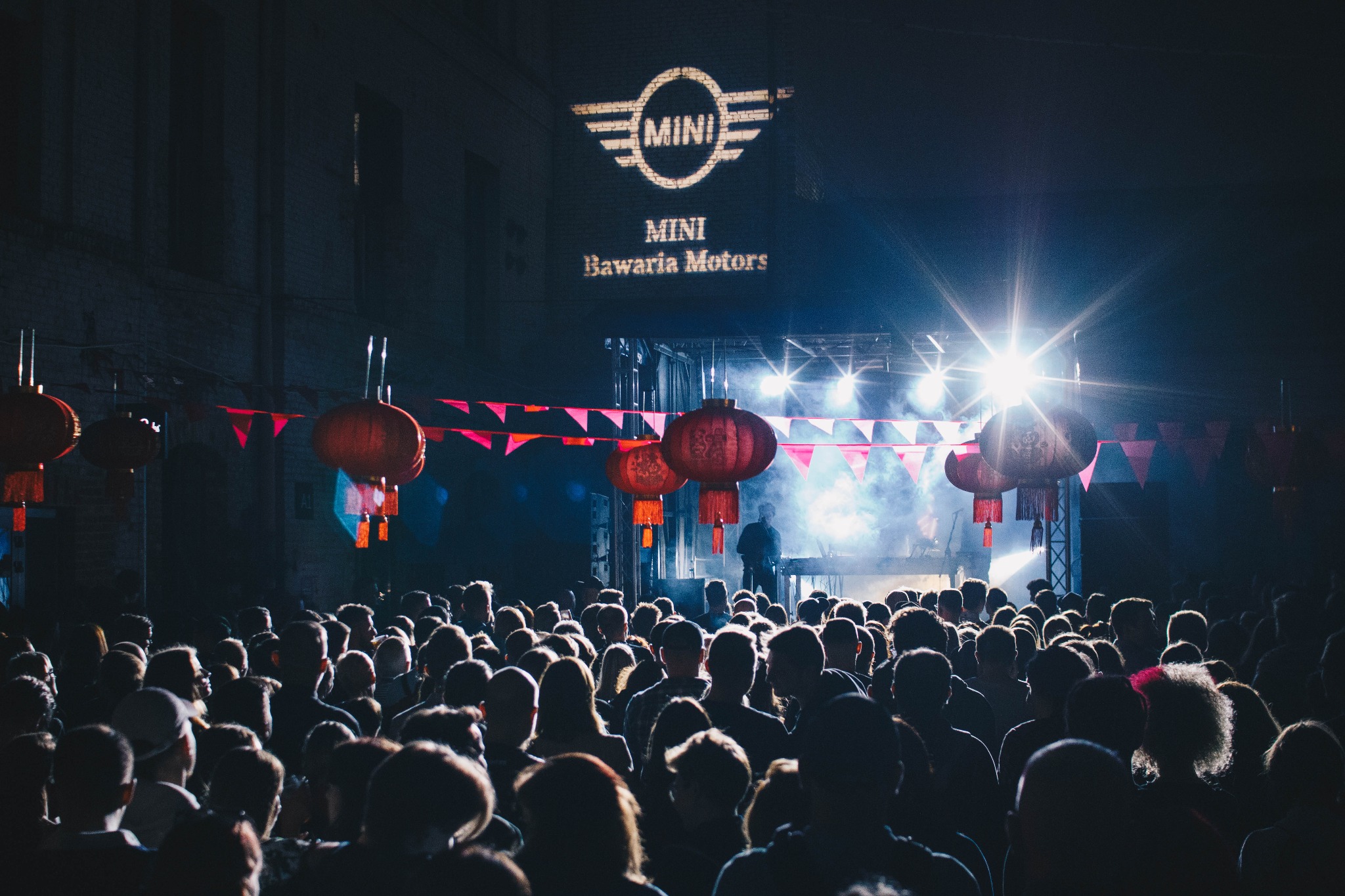 Tłum ludzi obserwuje scenę z oświetleniem i projekcją logo MINI na ścianie budynku podczas wydarzenia z dekoracjami w postaci czerwonych lampionów.