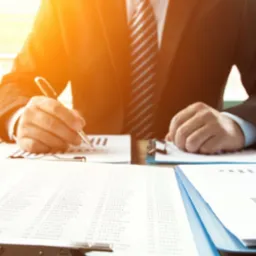 Mężczyzna w garniturze analizuje dane finansowe, wypełniając dokumenty na biurku z intensywnym, ciepłym światłem słonecznym wpadającym przez okno, podkreślającym profesjonalny charakter pracy.