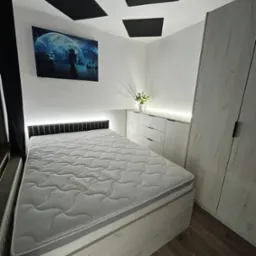 Nowoczesna sypialnia z ł&oacute;żkiem z podświetlanym zagł&oacute;wkiem, szafą z drewnopodobnymi frontami i komodą z trzema szufladami, całość w jasnej kolorystyce, na ścianie obraz z motywem żaglowc&oacute;w i księżyca