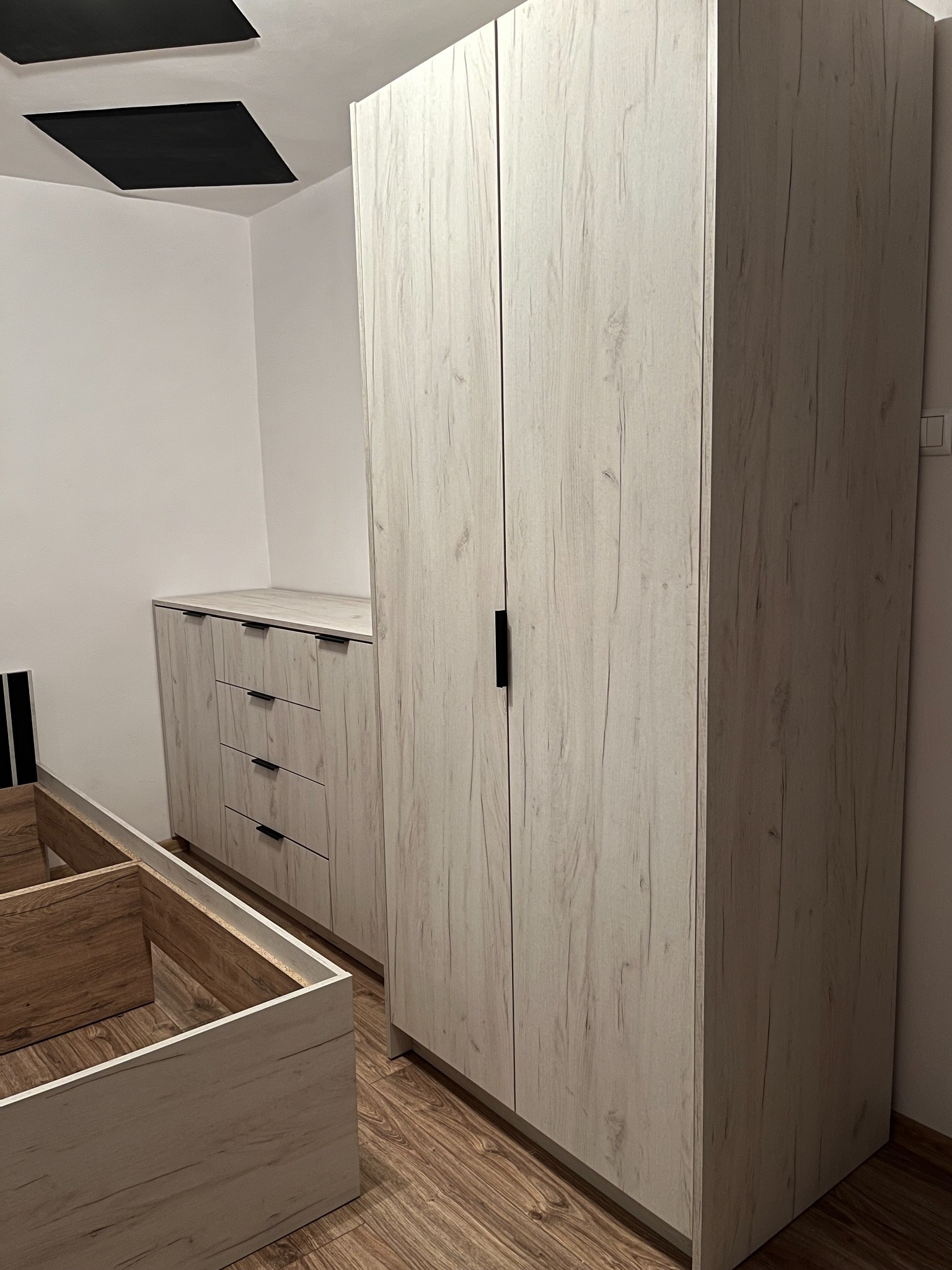 Jasna szafa dwudrzwiowa z czarnym uchwytem, komoda z szufladami i rama łóżka w pokoju z panelami na suficie.